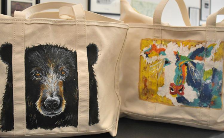 Megan Broome/The Clayton Tribune. The North Georgia Arts Guild “Original Art Treasure Tote” raffle features these 14-inch by 23-inch zippered L.L. Bean canvas totes containing over two dozen original, hand-made art treasures made by members of The North Georgia Arts Guild. The hand-painted exteriors feature a black bear by artist Penny Bradley (left) and a colorful cow by artist Kathy Beehler.