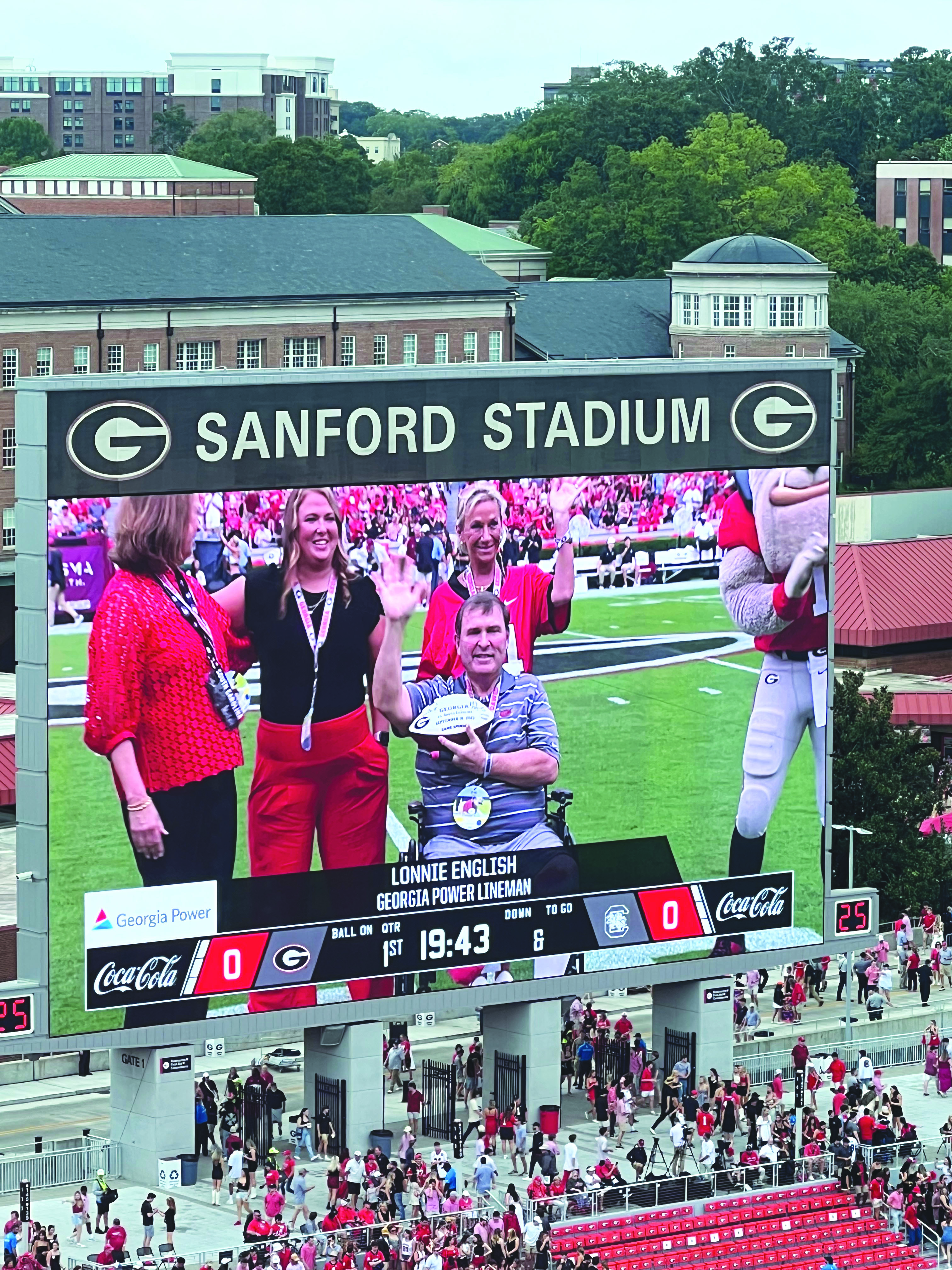 Submitted. Georgia Power lineman and Clayton resident Lonnie English, waves to over 96,000 fans while being honored at the Sept. 16 Georgia vs South Carolina football game after overcoming a traumatic leg injury. Joining English on the 50-yard line of Dooley Field at Sanford Stadium. Pictured from left to right is GA Power Chairman, President and CEO, Kim Greene, English’s daughter Logan, English’s wife Caroline and Georgia Mascot Hairy Dawg. 