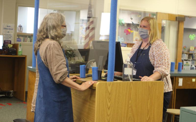 Megan Broome/The Clayton Tribune. Peggie Wilcox, circulation clerk, left, and Melissa Poslusny, senior clerk, demonstrate checkout procedures amidst the library's new contact precautions during the soft reopening of the Rabun County Library after being closed due to COVID-19.