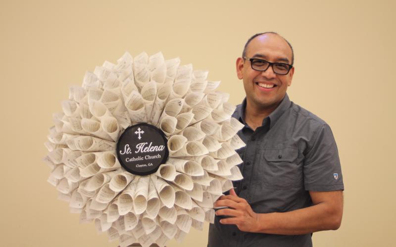 Megan Broome/The Clayton Tribune. Father Pedro Poloche holds up a wreath made by a parishioner at St. Helena Catholic Church. The wreath is made out of church bulletins that Poloche saved to write down the names of every family in the congregation to remember to pray for them during times of hardship. Each bulletin represents a different family name.