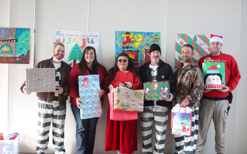 Megan Broome/The Clayton Tribune. Austin Groom, left, Captain Beth Darnell, Officer Patti Saunders, Joshua Arnold, Johnny Riddle and Officer Nathaniel Cross pass out Christmas gifts to the children of inmates on Saturday. They stand in front of the Christmas card made by inmates in C-Pod (center), who won the decorating contest.