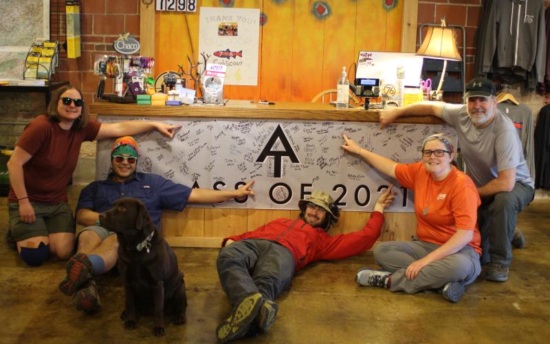 Megan Broome/The Clayton Tribune. Appalachian Trail (AT) hikers point to their trail names after signing the banner at Outdoor 76 on Tuesday. The group was being shuttled back to Dicks Creek Gap at the AT after staying overnight in Clayton. Each hiker has a unique name given to them by another hiker. Skylar Bounds (Chef), left, Nabal Camacho (Yeetnmouse), Jason Conn (70 lb Hammer), Kelsey Widener (Jingles) and Kip Beegle (Captain). Dog Telico also poses with the hikers.