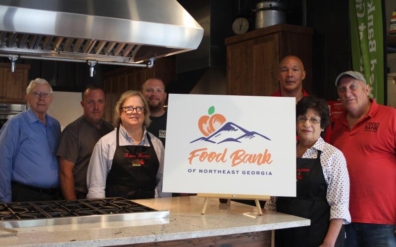 Megan Broome/The Clayton Tribune. Representatives from area food banks attended an unveiling of the new Food Bank of Northeast Georgia logo in the Food Bank Teaching Kitchen last week. Richard Boone, left, Aaron Holt, Laura Hunter, Philip Wilkes, James Brim, director of the Food Bank Mountain Branch, Joanna Baumgartner and Dave Smith stand in front of the new logo after it is revealed.