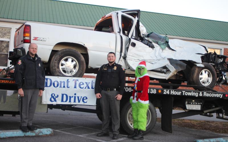 Megan Broome/The Clayton Tribune. Dillard Police Sgt. Michael Carnes, left, Chief Jeremy Parker and Sgt. Victoria Hopper (dressed as The Grinch) educate community members on traffic safety and consequences of distracted driving at the  “A Night Out in Christmastown” event held in Dillard Dec. 8. A sign reading “Don’t text and drive” was displayed before a wrecked vehicle sitting on a tow truck so community members could see visually potential consequences of distracted driving.