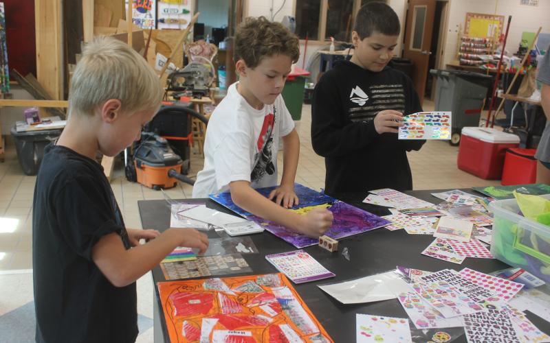 Megan Broome/The Clayton Tribune. Hayes Ward, 6, left, Ethan Rolader, 10, and Talon Stewart, 10, have sticker and coloring fun to finish their art pieces during Art Camp with Mrs. Amy Jarrard, Visual Arts teacher and Fine Arts Department Chair at Rabun County High School on July 14. Students participated in Art Camp several days last week and created many different works of art.