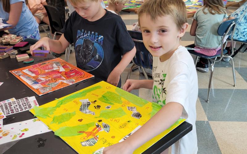 Megan Broome/The Clayton Tribune. Tagg Bedingfield, 6, has a huge smile as he shows off the art project he made during Art Camp with Mrs. Amy Jarrard last week. Pictured in the background are Hayes Ward, 6, and helper Abbye Carver.