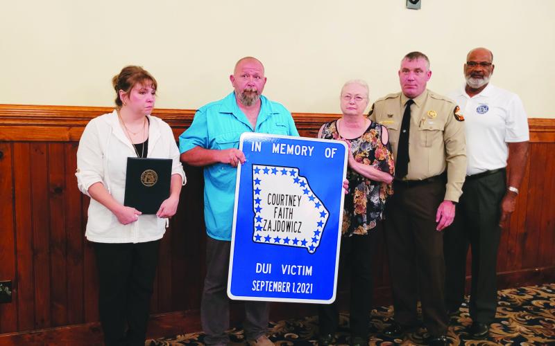 Photo by Megan Broome. Rabun County Sheriff Chad Nichols unveils a road sign honoring Rabun County Detention Center Officer Cpl. Courtney Faith Zajdowicz at the 31st annual Hands Across the Border event at The Dillard House Aug. 30. The sign will be erected at the crash site at George Eller Curve. Pictured are Courtney’s mother Crystal Zajdowicz, father David Cross, grandmother Janet Zajdowicz, Sheriff Chad Nichols and Allen Poole, director of the Georgia Governor’s Office of Highway Safety (GOHS).