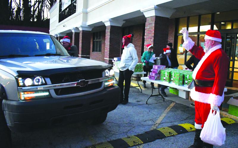 Enoch Autry/The Clayton Tribune. Santa waves to those in the long line at the Food Bank drive-thru to receive a wrapped gift. Wearing a seasonal hat (center), James Brim, the Mountain Branch director of the Food Bank of Northeast Georgia helps direct traffic.