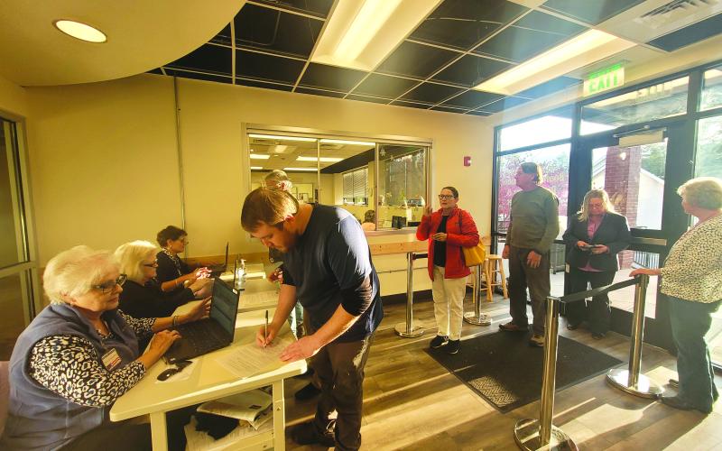 Megan Broome/The Clayton Tribune. The line of voters waiting to check in to cast their ballots extended outside of the elections office Tuesday afternoon 30 minutes before polls closed. In the forefront, poll worker Ann D’Agostino checks-in voter Caleb Watson as poll workers Sandra Wood and Kay Townsend also check in other voters. To the right, poll worker Jane Thomason helps assist voters through the process.