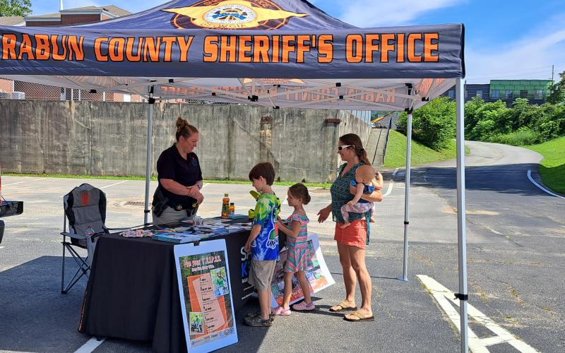 Megan Broome/The Clayton Tribune. School Resource Deputy Lisa Thompson with Safe Kids Rabun County provides community members important information about how to keep children safe using safety belts and helmets and information about the Georgia Passenger Safety Law, as well as other important information. During the Rabun County Health Department Diaper Day Resource Fair Saturday, July 8, Thompson talks with Samantha Grier and KarlieJae Grier, 7; Maxwell Grier, 10; and MaryAlice Grier, 7 months old.