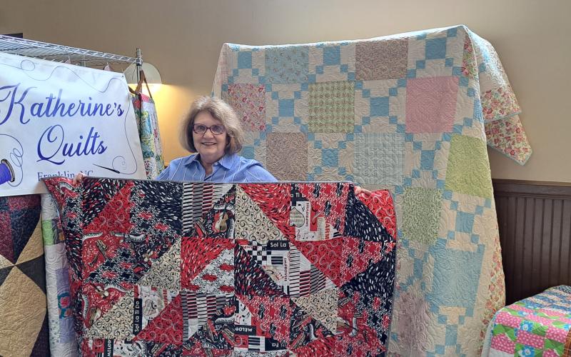 Megan Broome/The Clayton Tribune. Katherine Parker holds up a handmade quilt displaying music notes, script music and other music themes surrounded by “Katherine’s Quilts” during the Painted Fern Art Festival.