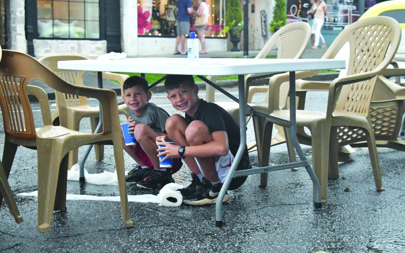 Enoch Autry/The Clayton Tribune. With their cups they got from a food truck, 11-year-old Grayson Miller and his brother Asher, 8, have a reason to smile as they stay dry under a table at the Downtown Clayton Block Party as a storm blew through Main Street on Aug. 12. Water droplets can be seen coming off the table as the paper towels next to their feet on the street are saturated.