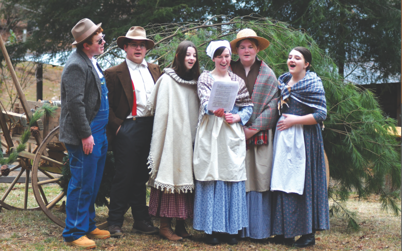 Enoch Autry/The Clayton Tribune. Rabun County High School students Isaac Craig, Andy Leff, Isabel Craine, Leizle Mason, Sarabeth Speed and Marlena Turpin sing holiday songs near the Warwoman Cabin and Phillips Cabin at the Dec. 16 annual “A Foxfire Christmas” in Mountain City. Left: Kim Cannon from the Rabun County Public Library read a variety of books that depicted pioneer life in the Appalachian Mountains on the Shooting Creek Cabin porch.