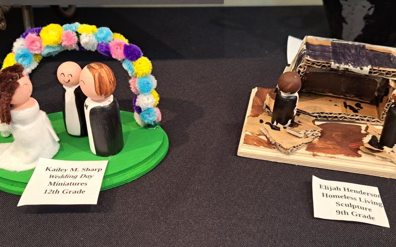 Megan Horn/The Clayton Tribune. Senior Kailey M. Sharp’s “Wedding Day” miniatures, left, and Ninth grader Elijah Henderson’s “Homeless Living” sculpture were on display during the RCMS and RCHS Art Showcase held in the Fine Arts Building (FAB) May 16. Left: RCHS senior Ryleigh Brotherton displays artwork in red clay titled “Friendly Miner."