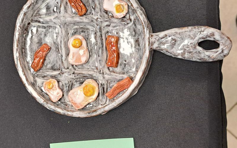 Megan Horn/The Clayton Tribune. Rabun County Middle School student Manny Martinez displayed this detailed work of art depicting eggs and bacon in a skillet during the RCMS and RCHS Art Showcase held in the Fine Arts Building (FAB) May 16. Left: RCHS senior Ryleigh Brotherton displays artwork in red clay titled “Friendly Miner."