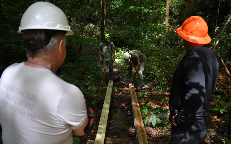 Enoch Autry/The Clayton Tribune. Brent Martin and Don Reis watch Scott Prim and Casey Quarterman work on a pedestrian trail bridge on May 10 as crews improved the walking trails at Warwoman Dell. Left: Volunteer Jim Chance chops trees in the Warwoman Dell area as part of the early morning cleanup. Others walked trails picking up trash.