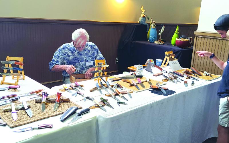 Megan Horn/The Clayton Tribune. Chris and Carson Moletz, right, enjoy viewing the detailed work of Jim Craig with his handmade knives during the 2024 Painted Fern Art Festival July 13 and 14.  Below: Submitted. At the 2024 Painted Fern Art Festival, Sean and Ebony Wilson (left) were the Producers’ Choice Award winners for Best Booth with their woodworking. Sharon Mumbi (right) was chosen by show visitors and awarded the Peoples’ Choice for Best in Show with her mixed fiber-leather, beads and fabric.