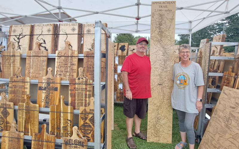 Megan Horn/The Clayton Tribune. Penny and Glenn Trotti with their business “PNG Custom Gifts” pose with their handcrafted works, using methods such as wood burning, during last weekend’s 27th Dillard Bluegrass Benefit Festival. This tall creation outlines the Appalachian Trail.