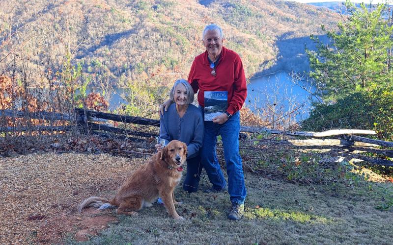 Megan Horn/The Clayton Tribune. A picture viewing a snowy mountain range and snow-covered fence at Lake Burton taken by Chip Goen of Clayton is now featured on the cover of the Winter 2024 Lake Living magazine. Lin Goen submitted the photograph to the contest. The Goens are pictured here with their dog Charley, the Lake Living magazine cover and $100, with the same backdrop in a different season.