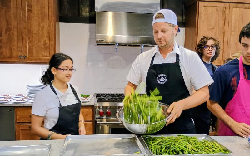 Megan Horn/The Clayton Tribune. Student Tara Shryock watches as Chef Ryan Miller, culinary and partnerships coordinator for the Food Bank of Northeast Georgia Mountain Branch, tosses a bowl of asparagus for a four-course meal served at the Teaching Kitchen on July 25. The meal was the culmination of three days of an Advanced Cooking Class. Also pictured are Tessa Stuckey and Hank Stuckey.