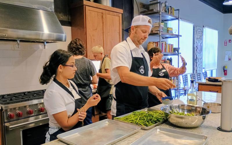 Megan Horn/The Clayton Tribune. Student Tara Shryock watches as Chef Ryan Miller, culinary and partnerships coordinator for the Food Bank of Northeast Georgia Mountain Branch, prepares asparagus for a four-course meal served at the Teaching Kitchen on July 25. Pictured in the background preparing food are students Tessa Stuckey, Brantley Lavender and Kitchenettes Volunteer Joanne Baumgartner.