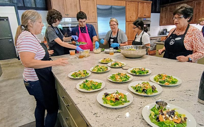 Megan Horn/The Clayton Tribune. Kitchenettes Volunteer Laura Josephson; Tessa Stuckey; Hank Stuckey; Kitchenettes Volunteer Annette Branan; Tara Shryock; and Kitchenettes Volunteer Joanne Baumgartner, prepare an Irish flag salad with homemade dressing during the July 25 dinner at the Teaching Kitchen. A four-course meal prepared by students culminated three days of an Advance Cooking Class as part of the Teaching Kitchen program.