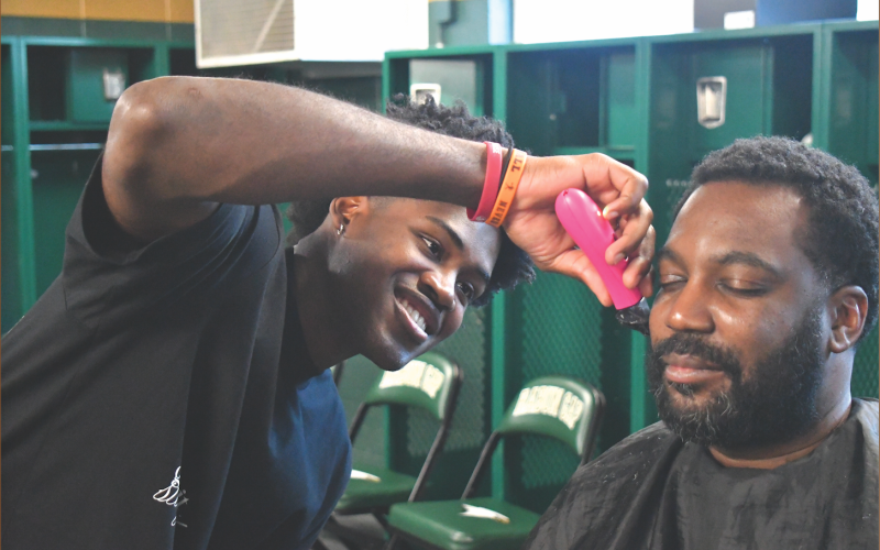 Wesley Branch/The Clayton Tribune. A smiling Rabun Gap senior RJ Lacossade from Turks and Caicos cuts Rabun Gap boys assistant basketball coach Darrick Cureton’s hair in the Rabun Gap-Nacoochee School locker room on Jan. 12. Lacossade has been cutting hair for several years now, cutting his own hair and other students at RGNS.