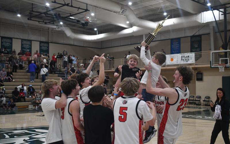 Wesley Branch/The Clayton Tribune. The Rabun County boys basketball team celebrates after being awarded the championship trophy for winning the Region 8 Tournament of Class A Division I, following its 58-53 overtime win over Commerce on Feb. 20 at Piedmont University. Hoisting the trophy while being lifted by senior forward Hayes Free is Reeves Page, the son of assistant coach Corey Page.
