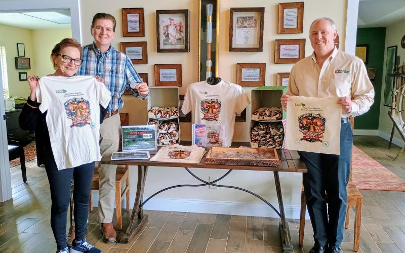 Megan Horn/The Clayton Tribune. The Rabun County Welcome Center has plenty of swag for sale as part of America’s 250th birthday celebration including challenge coins commemorating 250 years, canvas tote bags and t-shirts. Pictured holding the merchandise are Welcome Center Manager Linda Scofield, left; Mickey Duvall, business and economic development strategist; and Rick Story, Rick Story, president of Forward Rabun/Rabun County Chamber of Commerce.