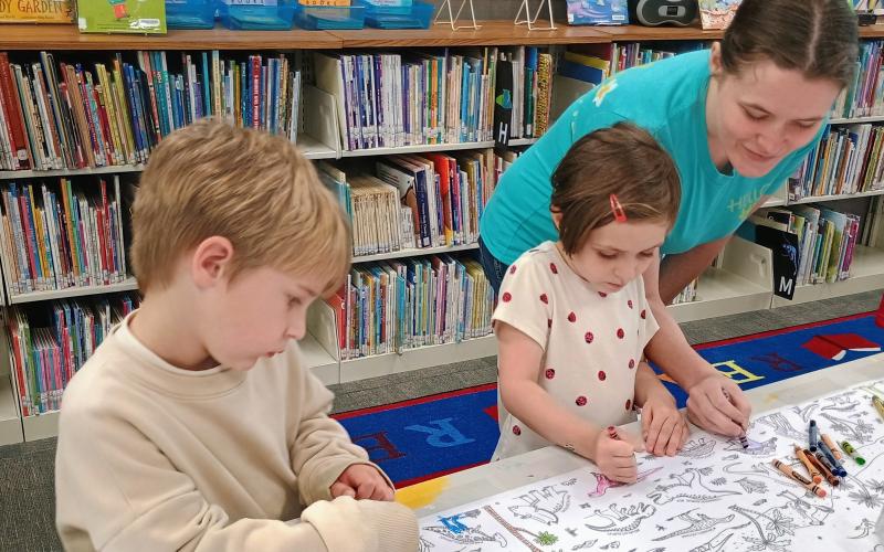 Megan Horn/The Clayton Tribune. Ollie Hightower, 4, Cora Brown, 4, enjoy coloring pictures of dinosaurs during the June 2 summer reading kick-off with the theme “Unearth a Story.” Also pictured is Katie Tench.