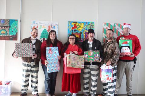 Megan Broome/The Clayton Tribune. Austin Groom, left, Captain Beth Darnell, Officer Patti Saunders, Joshua Arnold, Johnny Riddle and Officer Nathaniel Cross pass out Christmas gifts to the children of inmates on Saturday. They stand in front of the Christmas card made by inmates in C-Pod (center), who won the decorating contest. 
