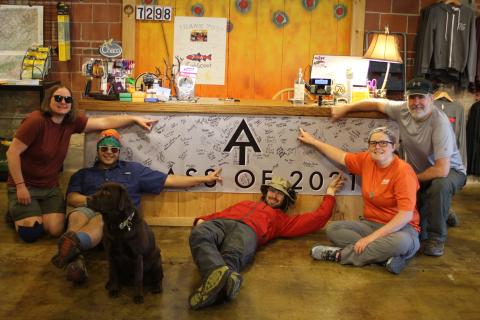 Megan Broome/The Clayton Tribune. Appalachian Trail (AT) hikers point to their trail names after signing the banner at Outdoor 76 on Tuesday. The group was being shuttled back to Dicks Creek Gap at the AT after staying overnight in Clayton. Each hiker has a unique name given to them by another hiker. Skylar Bounds (Chef), left, Nabal Camacho (Yeetnmouse), Jason Conn (70 lb Hammer), Kelsey Widener (Jingles) and Kip Beegle (Captain). Dog Telico also poses with the hikers. 