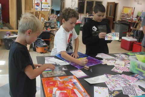 Megan Broome/The Clayton Tribune. Hayes Ward, 6, left, Ethan Rolader, 10, and Talon Stewart, 10, have sticker and coloring fun to finish their art pieces during Art Camp with Mrs. Amy Jarrard, Visual Arts teacher and Fine Arts Department Chair at Rabun County High School on July 14. Students participated in Art Camp several days last week and created many different works of art.  