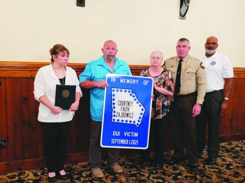 Photo by Megan Broome. Rabun County Sheriff Chad Nichols unveils a road sign honoring Rabun County Detention Center Officer Cpl. Courtney Faith Zajdowicz at the 31st annual Hands Across the Border event at The Dillard House Aug. 30. The sign will be erected at the crash site at George Eller Curve. Pictured are Courtney’s mother Crystal Zajdowicz, father David Cross, grandmother Janet Zajdowicz, Sheriff Chad Nichols and Allen Poole, director of the Georgia Governor’s Office of Highway Safety (GOHS). 