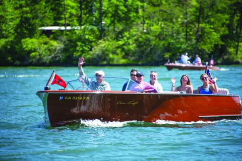 Submitted photo. A boat owner waves to the crowd during a previous LBCA Wooden Boat Parade show on Lake Burton. Left: Photo courtesy Conner Horn.  Rabun County is the Wooden Boat Capital of Georgia and Lake Burton Wooden Boat owners hold up the sign at the May 12 Captain’s reception. 