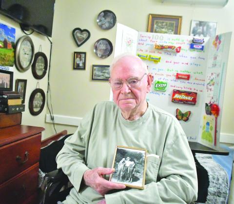Megan Broome/The Clayton Tribune. Ervin Chastain enjoys smiling, laughing and telling stories of his adventures in life. He was born on June 10, 1923, and recently celebrated the milestone with family, friends and former coworkers at Battle Branch Baptist Church. Pictured here, he holds a photo of himself at age 20 or 21 with parents Mack and Rosie Chastain. Behind him is a poster made by family members to honor his 100 years. 