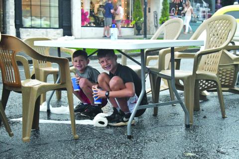 Enoch Autry/The Clayton Tribune. With their cups they got from a food truck, 11-year-old Grayson Miller and his brother Asher, 8, have a reason to smile as they stay dry under a table at the Downtown Clayton Block Party as a storm blew through Main Street on Aug. 12. Water droplets can be seen coming off the table as the paper towels next to their feet on the street are saturated. 