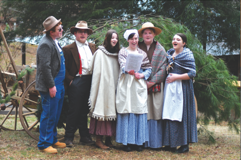Enoch Autry/The Clayton Tribune. Rabun County High School students Isaac Craig, Andy Leff, Isabel Craine, Leizle Mason, Sarabeth Speed and Marlena Turpin sing holiday songs near the Warwoman Cabin and Phillips Cabin at the Dec. 16 annual “A Foxfire Christmas” in Mountain City. Left: Kim Cannon from the Rabun County Public Library read a variety of books that depicted pioneer life in the Appalachian Mountains on the Shooting Creek Cabin porch.