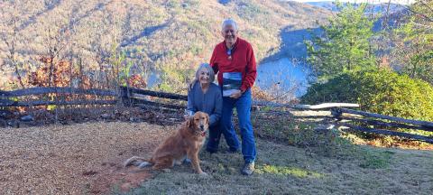 Megan Horn/The Clayton Tribune. A picture viewing a snowy mountain range and snow-covered fence at Lake Burton taken by Chip Goen of Clayton is now featured on the cover of the Winter 2024 Lake Living magazine. Lin Goen submitted the photograph to the contest. The Goens are pictured here with their dog Charley, the Lake Living magazine cover and $100, with the same backdrop in a different season. 