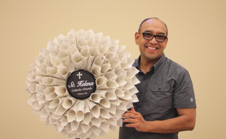 Megan Broome/The Clayton Tribune. Father Pedro Poloche holds up a wreath made by a parishioner at St. Helena Catholic Church. The wreath is made out of church bulletins that Poloche saved to write down the names of every family in the congregation to remember to pray for them during times of hardship. Each bulletin represents a different family name.