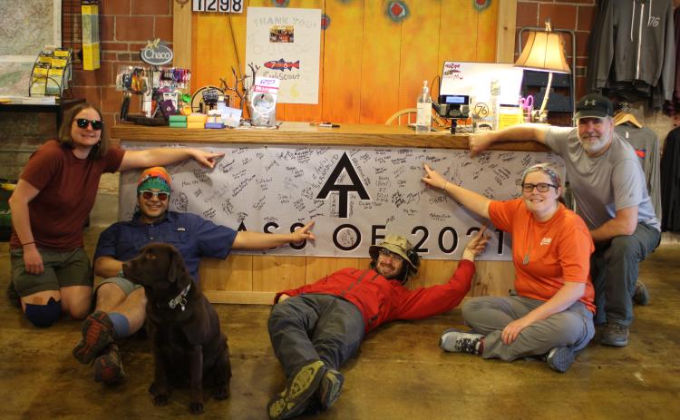 Megan Broome/The Clayton Tribune. Appalachian Trail (AT) hikers point to their trail names after signing the banner at Outdoor 76 on Tuesday. The group was being shuttled back to Dicks Creek Gap at the AT after staying overnight in Clayton. Each hiker has a unique name given to them by another hiker. Skylar Bounds (Chef), left, Nabal Camacho (Yeetnmouse), Jason Conn (70 lb Hammer), Kelsey Widener (Jingles) and Kip Beegle (Captain). Dog Telico also poses with the hikers.