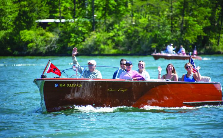 Submitted photo. A boat owner waves to the crowd during a previous LBCA Wooden Boat Parade show on Lake Burton. Left: Photo courtesy Conner Horn.  Rabun County is the Wooden Boat Capital of Georgia and Lake Burton Wooden Boat owners hold up the sign at the May 12 Captain’s reception.
