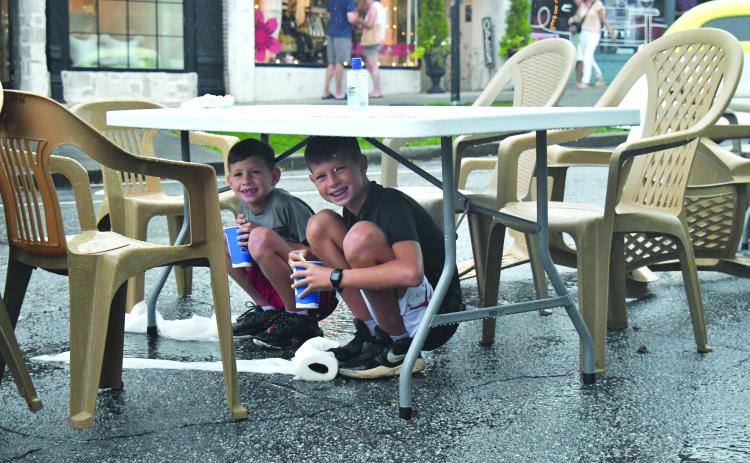 Enoch Autry/The Clayton Tribune. With their cups they got from a food truck, 11-year-old Grayson Miller and his brother Asher, 8, have a reason to smile as they stay dry under a table at the Downtown Clayton Block Party as a storm blew through Main Street on Aug. 12. Water droplets can be seen coming off the table as the paper towels next to their feet on the street are saturated.
