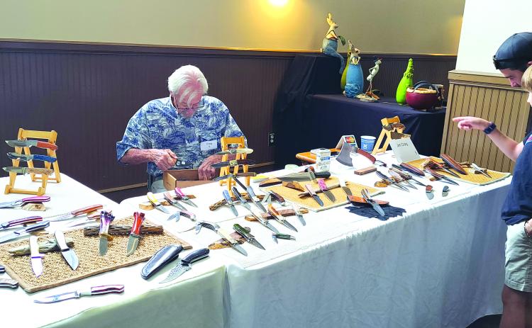 Megan Horn/The Clayton Tribune. Chris and Carson Moletz, right, enjoy viewing the detailed work of Jim Craig with his handmade knives during the 2024 Painted Fern Art Festival July 13 and 14.  Below: Submitted. At the 2024 Painted Fern Art Festival, Sean and Ebony Wilson (left) were the Producers’ Choice Award winners for Best Booth with their woodworking. Sharon Mumbi (right) was chosen by show visitors and awarded the Peoples’ Choice for Best in Show with her mixed fiber-leather, beads and fabric.