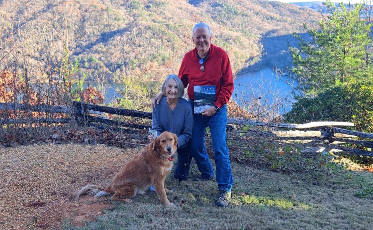 Megan Horn/The Clayton Tribune. A picture viewing a snowy mountain range and snow-covered fence at Lake Burton taken by Chip Goen of Clayton is now featured on the cover of the Winter 2024 Lake Living magazine. Lin Goen submitted the photograph to the contest. The Goens are pictured here with their dog Charley, the Lake Living magazine cover and $100, with the same backdrop in a different season.