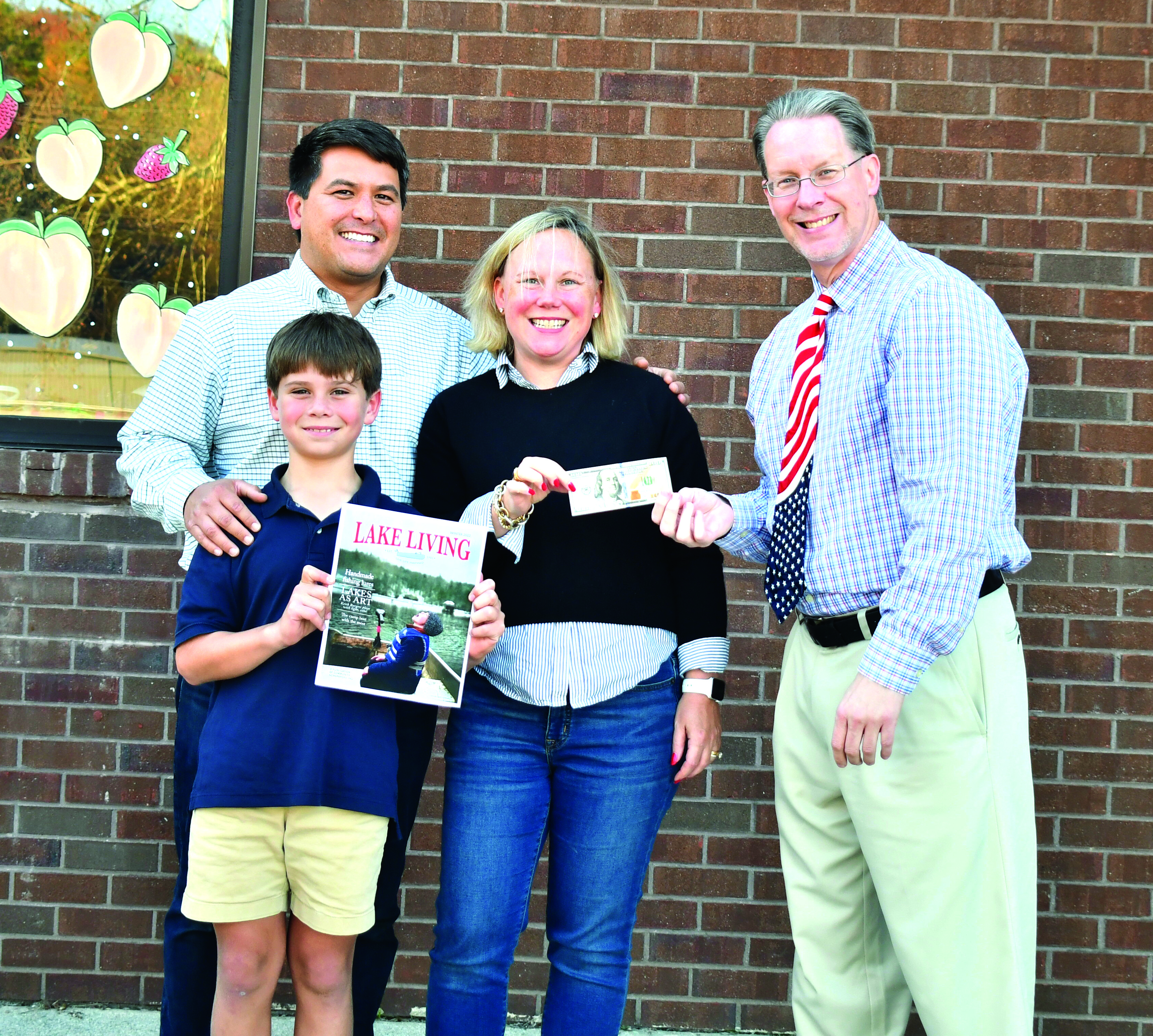 Megan Broome/The Clayton Tribune. Jonathon, Sarah, and Jonathon Barge Jr., who goes by “J,”  are all smiles as they learn that Jonathon Barge won the Winter 2022 Lake Living cover contest for the picture of “J” in the family’s 1972 Alumacraft fishing boat “catching snowflakes instead of fish” on Lake Burton in January 2022. Enoch Autry, publisher of The Clayton Tribune, hands the family the $100 prize for winning the contest. 