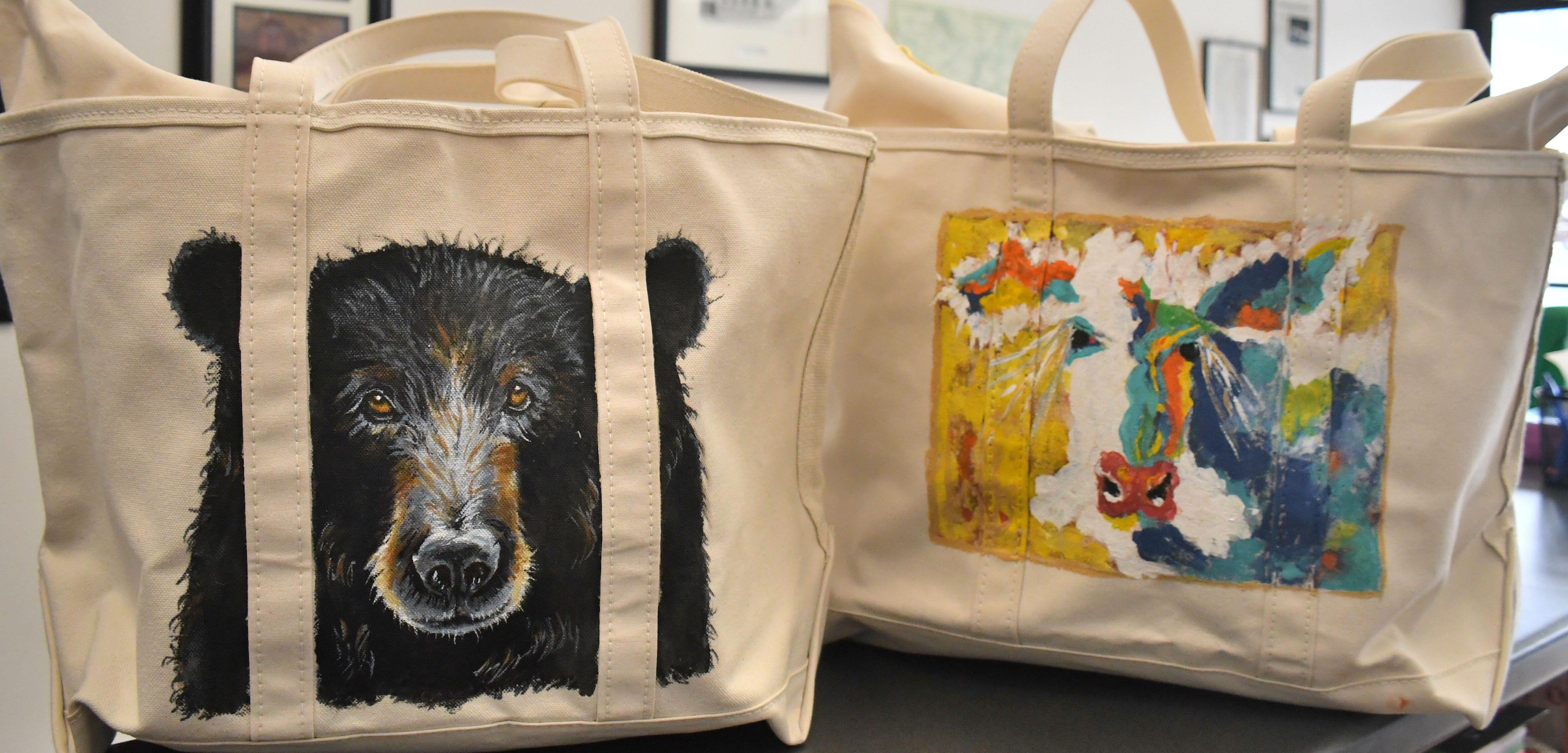 Megan Broome/The Clayton Tribune. The North Georgia Arts Guild “Original Art Treasure Tote” raffle features these 14-inch by 23-inch zippered L.L. Bean canvas totes containing over two dozen original, hand-made art treasures made by members of The North Georgia Arts Guild. The hand-painted exteriors feature a black bear by artist Penny Bradley (left) and a colorful cow by artist Kathy Beehler.