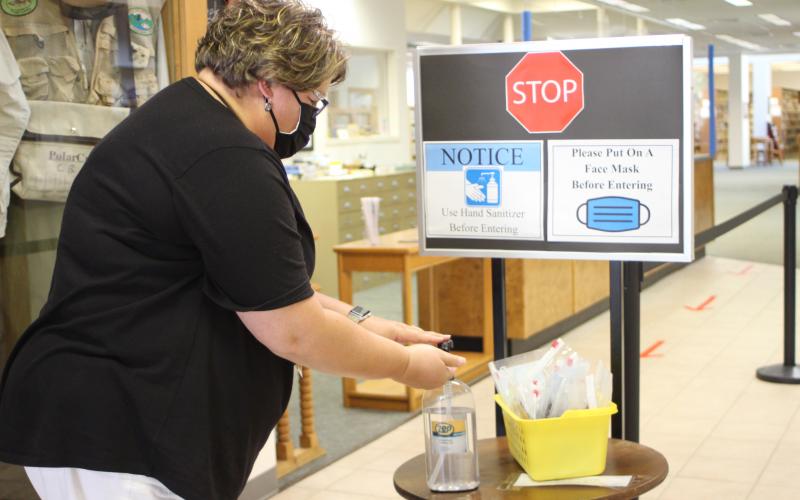 Megan Broome/The Clayton Tribune. The Rabun County Public Library reopened on Tuesday and patrons are required to wear masks while browsing and follow other health guidelines like sanitizing hands and following the direction arrows to maintain a six-foot distance.  Jennifer Buchanan, circulation supervisor, uses hand sanitizer to demonstrate measures patrons are asked to take upon entering the library. 