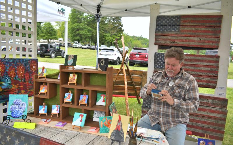 Megan Broome/The Clayton Tribune. Artist Jack Blades with WoodFlagArtStudio paints and displays his artwork at the inaugural Dinglerfest Saturday, May 6. His artwork features animals and plants and he also creates rustic vintage wood American flags.