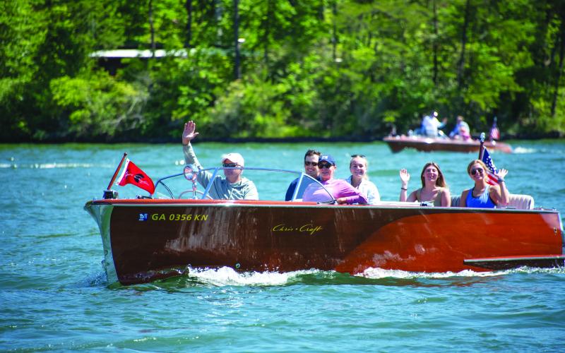 Submitted photo. A boat owner waves to the crowd during a previous LBCA Wooden Boat Parade show on Lake Burton. Left: Photo courtesy Conner Horn.  Rabun County is the Wooden Boat Capital of Georgia and Lake Burton Wooden Boat owners hold up the sign at the May 12 Captain’s reception. 